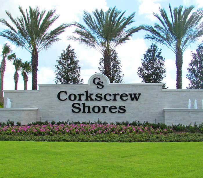 Entrance sign for the Corkscrew Shores community, located off Corkscrew Road in Southwest Florida, with palm trees, lush greenery, and colorful flower beds in the background.