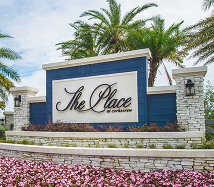Blue and white entrance sign for "The Place at Corkscrew," a welcoming community on Corkscrew Road, surrounded by palm trees and pink flowers.