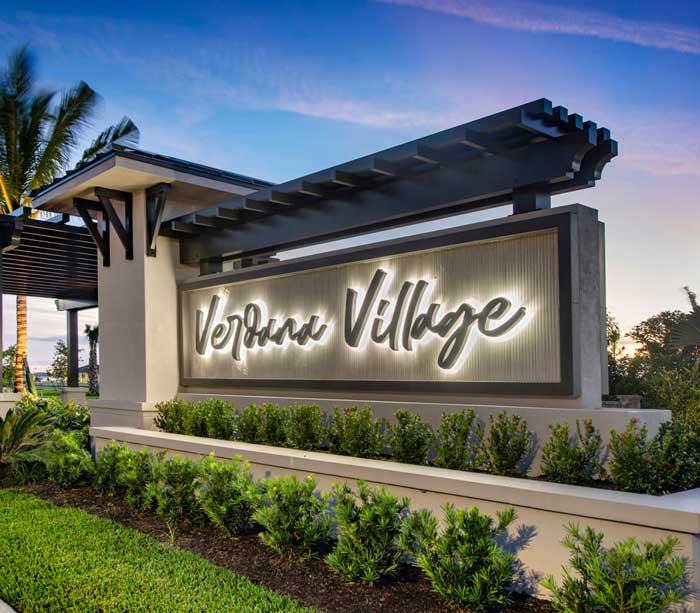 A modern outdoor sign with illuminated cursive lettering reads "Veranda Village," welcoming the community along Corkscrew Road, surrounded by lush landscaping and set against a Southwest Florida dusk sky.